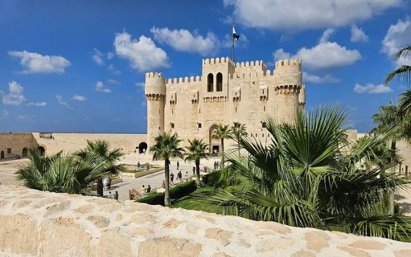 Qaitbay Fort in Egypt under a bright blue sky with palm trees in front.