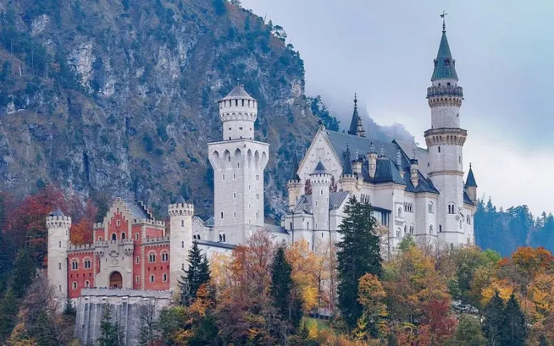 Neuschwanstein Castle in Germany rises dramatically against forested cliffs in autumn.