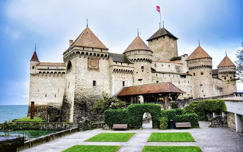 Chillon Castle in Switzerland with round towers beside a lake and green hedges in front.