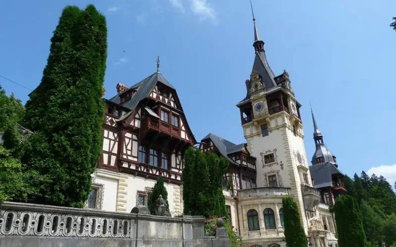 Peleș Castle in Romania with ornate towers, timber framing, and tall green trees in front.