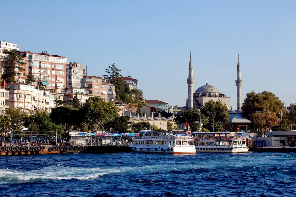 Waterfront view of ferries at a pier with the Mihrimah Sultan Mosque and hillside homes under a clear sky