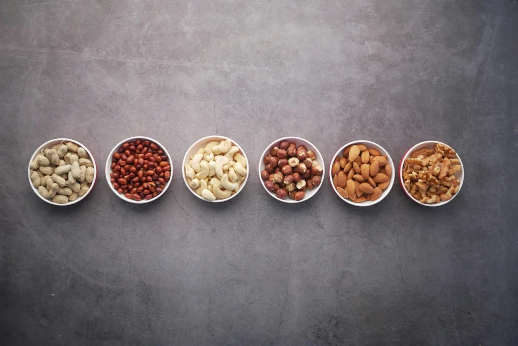 Six small bowls of different nuts lined up horizontally on a grey surface