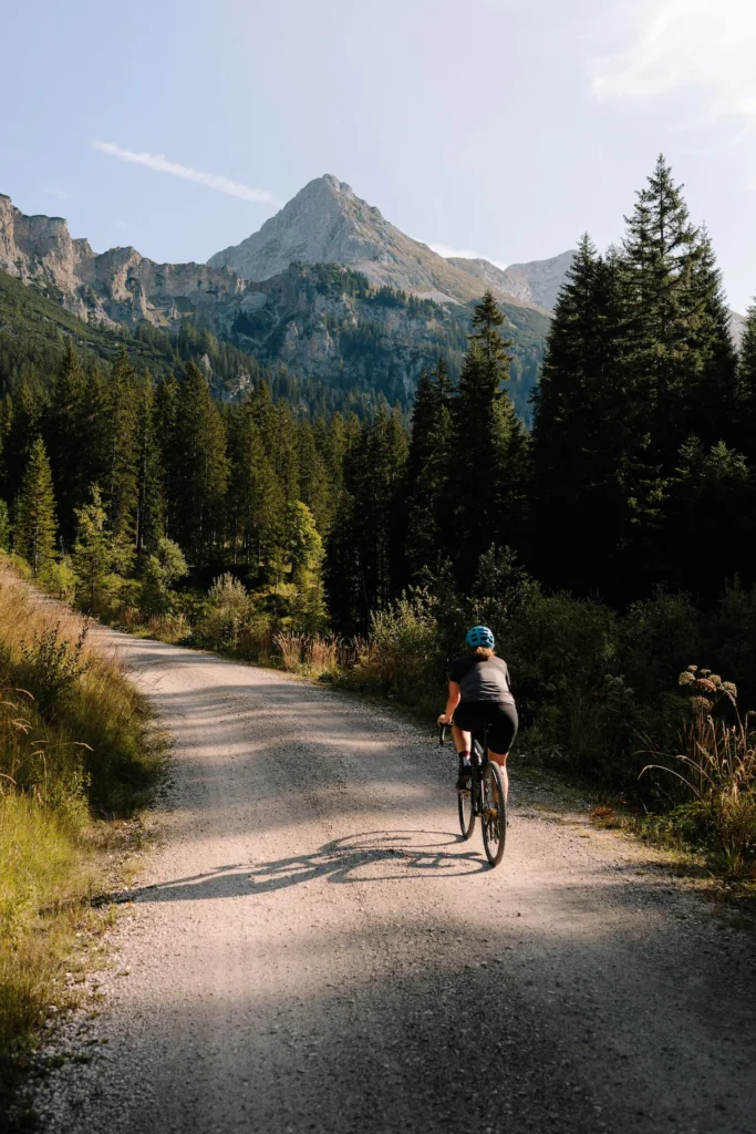Cyclist riding a gravel bike up a dirt path toward a rocky mountain peak lined with pine trees