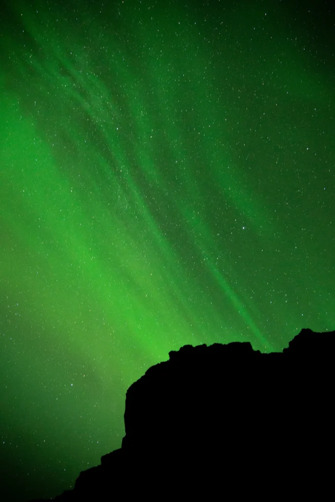 Vibrant green aurora borealis streaks filling a starry night sky above a dark mountain silhouette