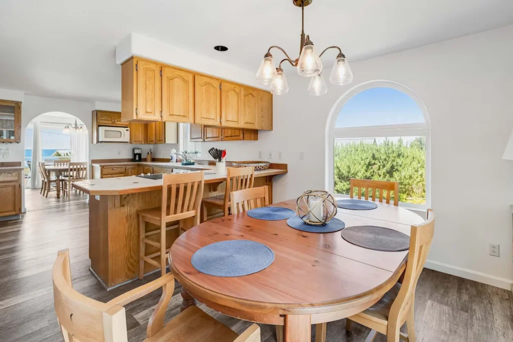 Beach house dining area with a wooden table, kitchen island barstools, and an arched window looking out at coastal greenery