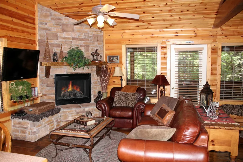 A cozy cabin living room with pine wood walls, a stone fireplace, brown leather furniture, and a ceiling fan