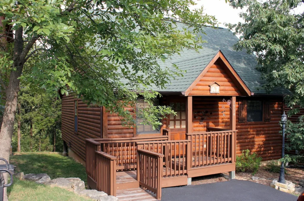 A rustic two-story log cabin with a green shingled roof, a wooden front porch, and an accessible ramp surrounded by trees