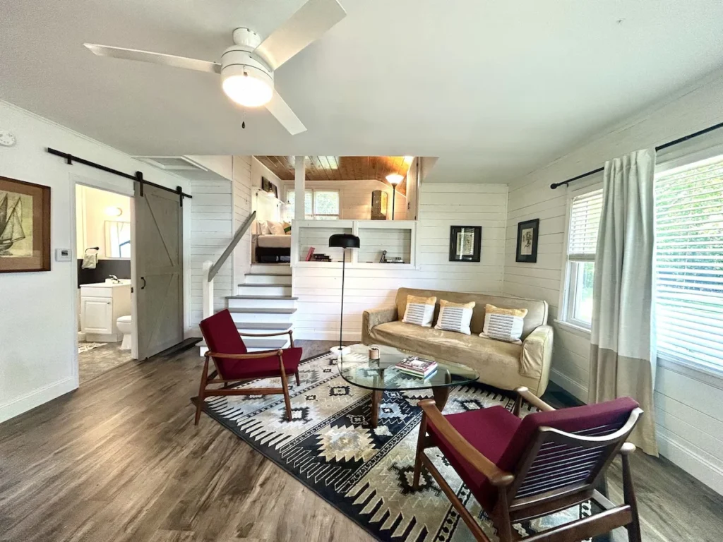 Bright cottage living room with a patterned rug, two maroon armchairs, a tan sofa, and stairs leading up to a loft