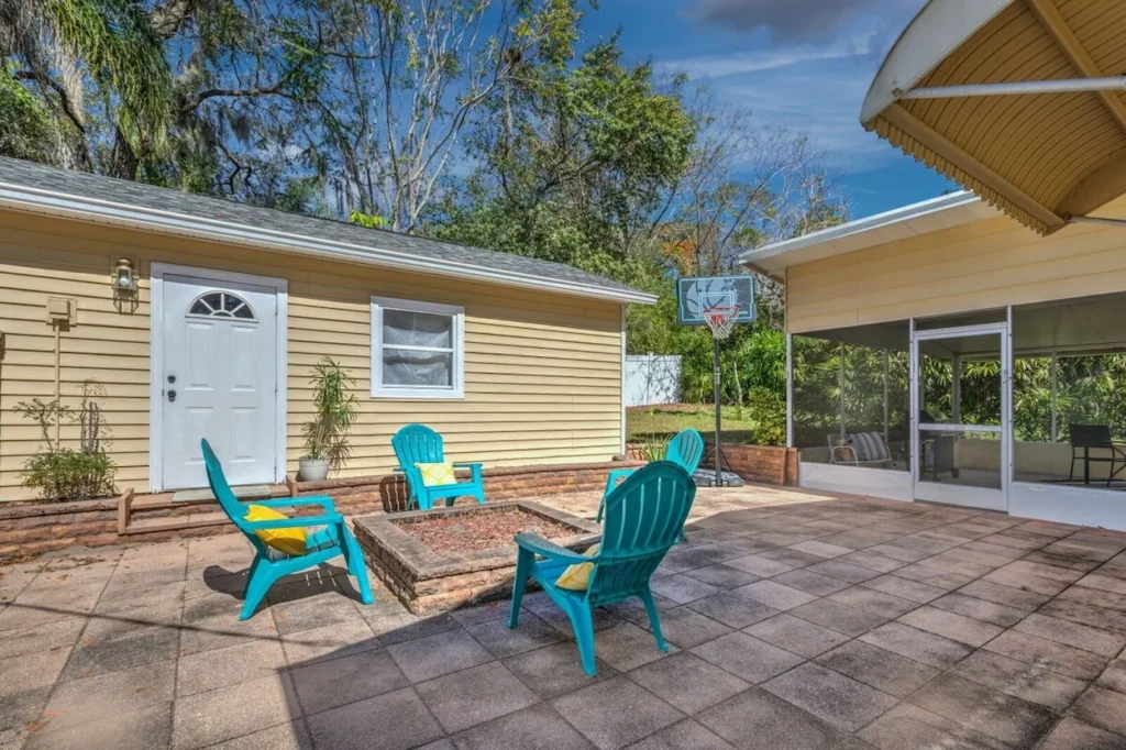Paved backyard patio with a central stone fire pit, teal Adirondack chairs, a detached yellow shed, and a basketball hoop