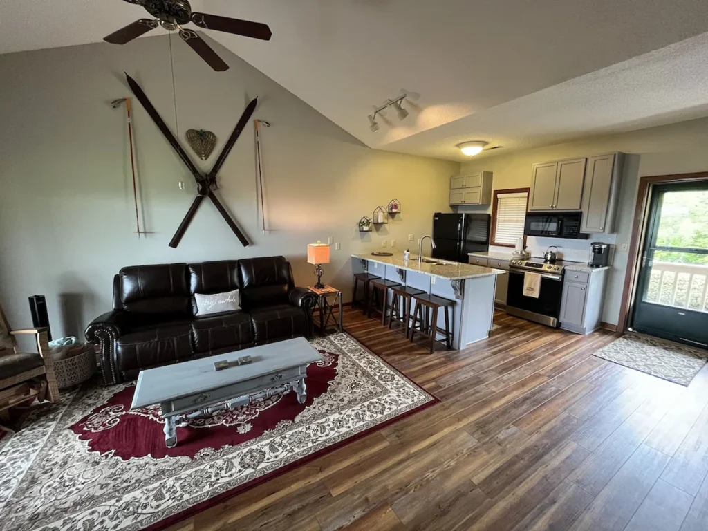 Open-concept cabin interior with a brown leather sofa below crossed wall skis, a central island, and grey kitchen cabinets