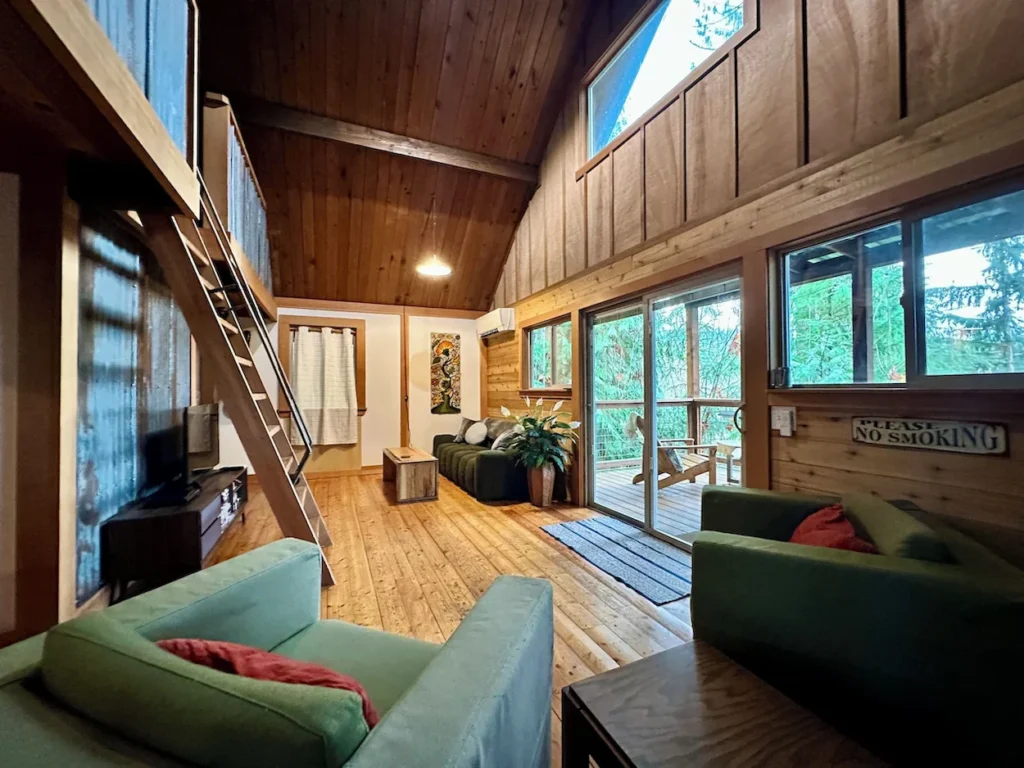 A rustic cabin living room with green armchairs, a wooden ladder to a loft, and glass sliding doors leading to a deck