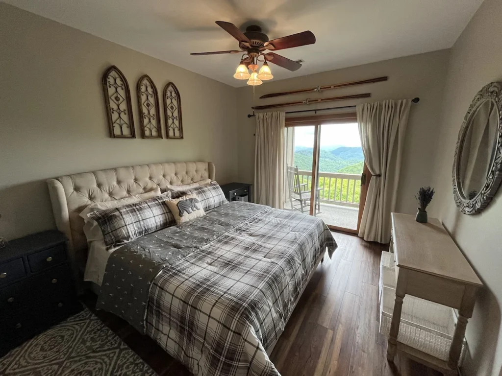 A bedroom with a tufted headboard, plaid bedding, and sliding glass doors opening to a scenic mountain balcony