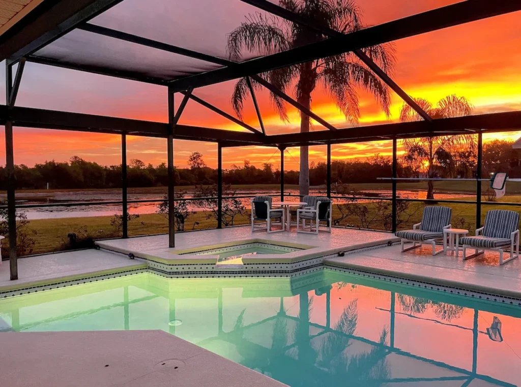 An enclosed backyard swimming pool and hot tub overlooking a lake under a vibrant orange and red sunset