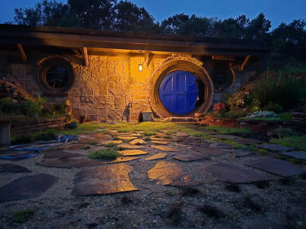 An earthen hobbit house with stone walls and a bright round blue door illuminated by a warm outdoor light at dusk