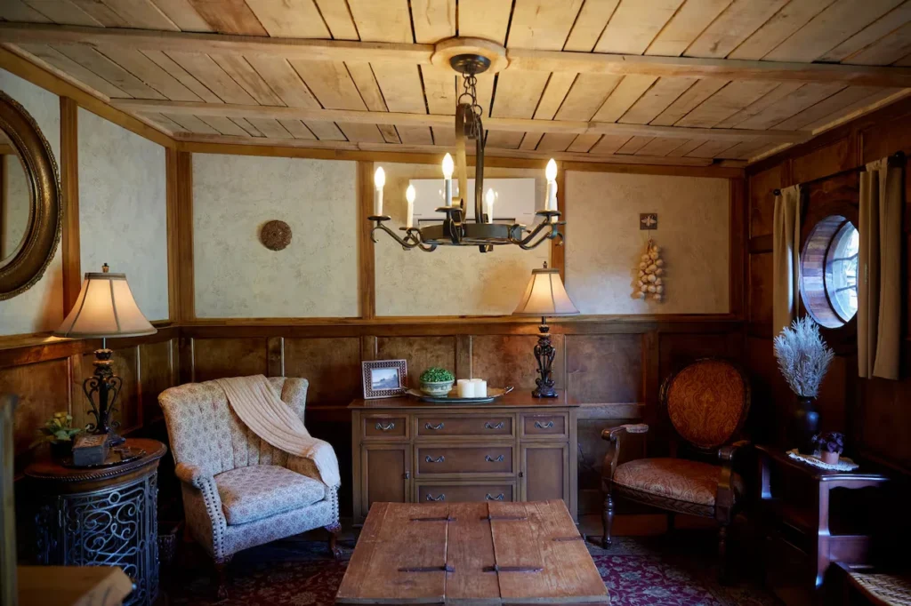 A rustic sitting room with wood-paneled walls, two patterned armchairs, a central chandelier, and a small round window
