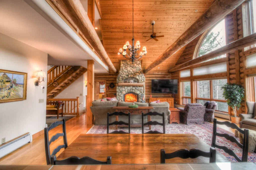 Log cabin interior looking over a wooden dining table toward a stone fireplace and vaulted wood ceilings