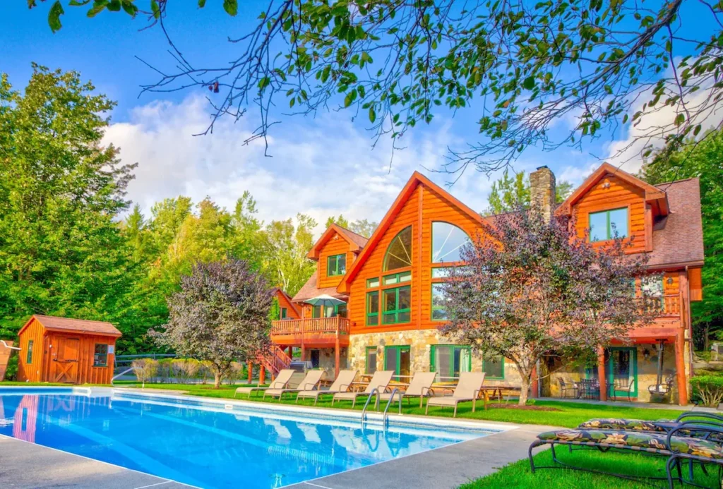 A large timber log house with stone accents, a stone chimney, and an outdoor swimming pool lined with lounge chairs