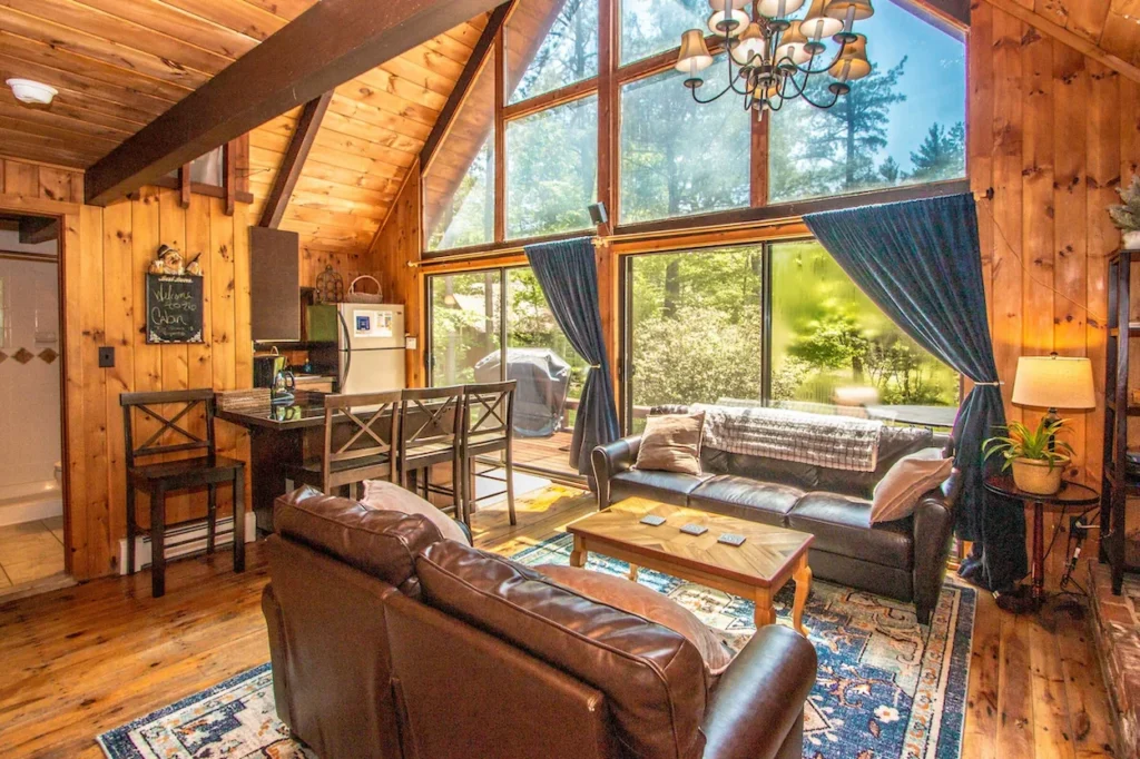 Rustic wooden cabin living room with towering glass windows, leather couches, and a small kitchen breakfast bar
