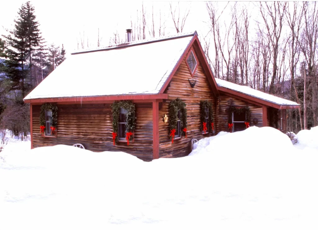 Rustic log cabin covered in deep snow with green holiday wreaths hanging on the windows