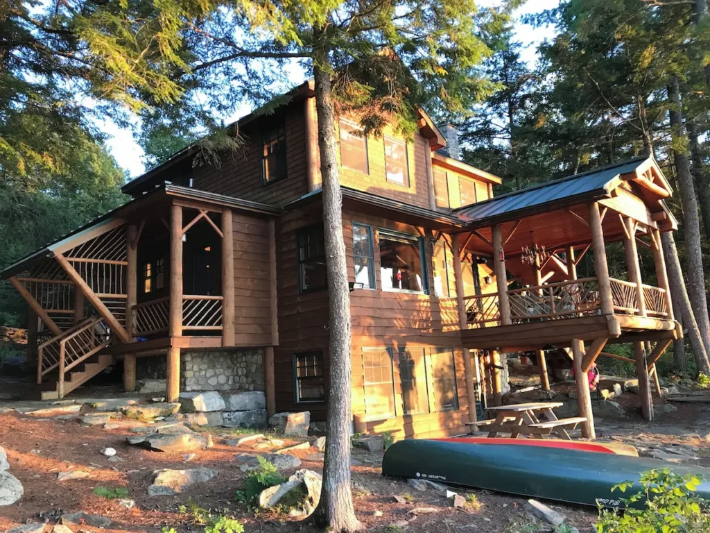 Large multi-story wooden log cabin in a pine forest with a green canoe resting on the rocky ground out front