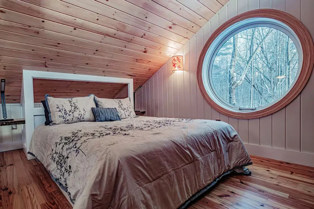 Cabin loft bedroom with a slanted wood ceiling, a queen-sized bed, and a large round portal window looking out at trees