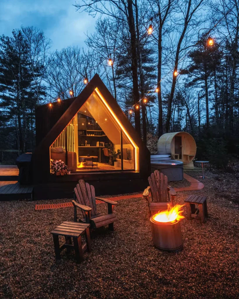 Illuminated A-frame cabin at dusk with an outdoor fire pit, hot tub, and wood barrel sauna