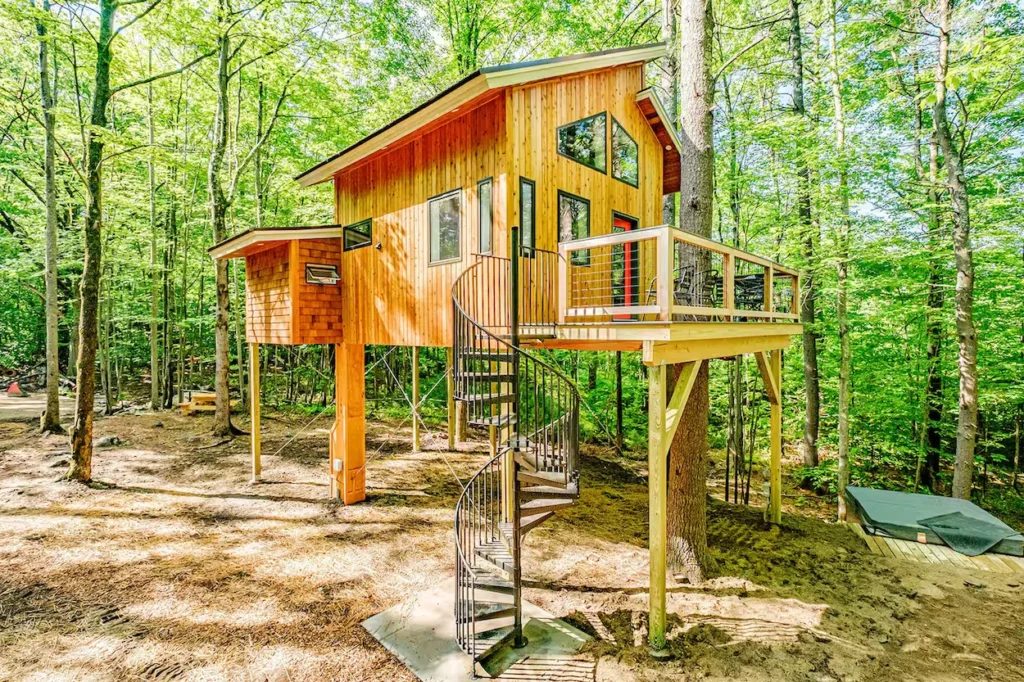 Modern wooden treehouse elevated on stilts with a black spiral staircase leading to an outdoor deck