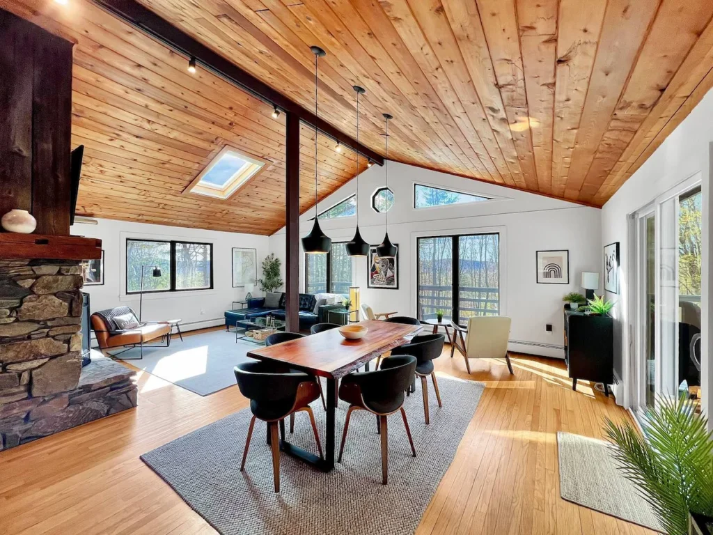Open concept dining area under a slanted wood ceiling with black pendant lights and a stone fireplace