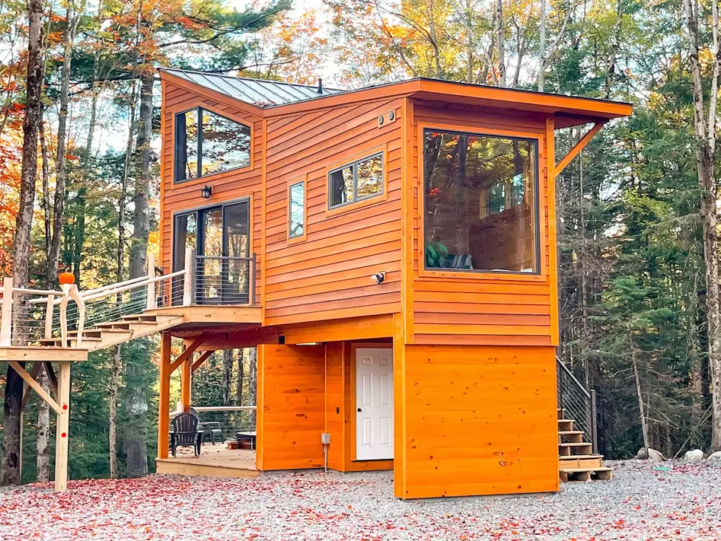 Modern two-story orange wood cabin elevated on stilts with a raised walkway surrounded by autumn trees