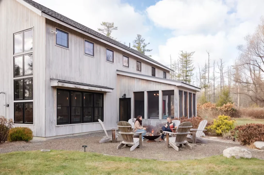 Two people sitting in wooden Adirondack chairs around a fire pit outside a modern vertical siding farmhouse
