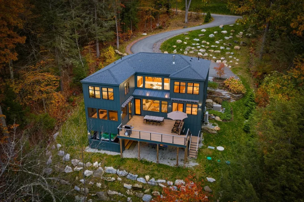Aerial view of a modern two-story dark green house with a large deck surrounded by autumn trees
