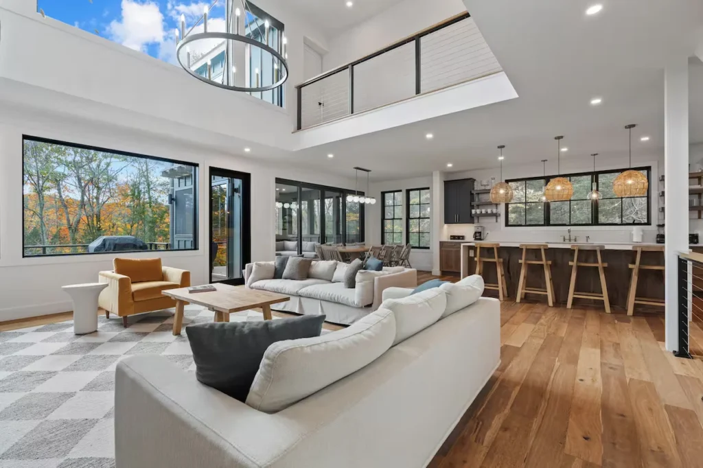 Modern open-concept living area with white walls, a loft balcony, large windows, and a kitchen island with wooden barstools