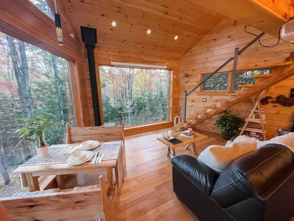 Wood-paneled cabin interior featuring a dining table, leather sofa, and a massive glass window showcasing autumn trees
