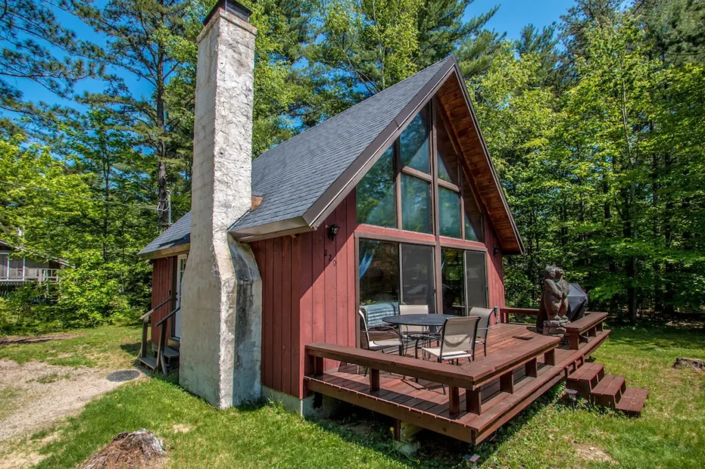 Red wood A-frame cabin with a large stone chimney and an outdoor wooden deck with a patio table
