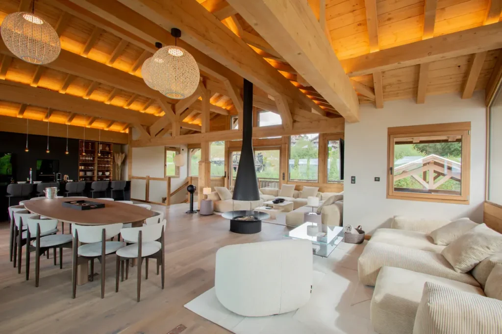 Chalet L'Ours Blanc interior featuring a central hanging fireplace, exposed timber beams, and a large dining table