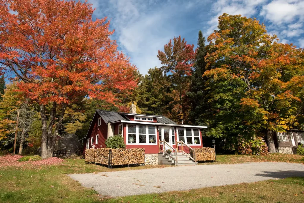 Small red cottage with white trim and stacked firewood piles surrounded by colorful fall trees