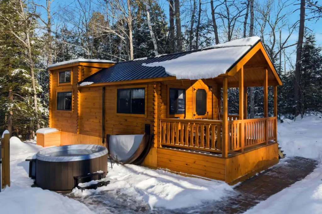 Small wooden cabin with a snow-covered roof and an outdoor hot tub in the snow