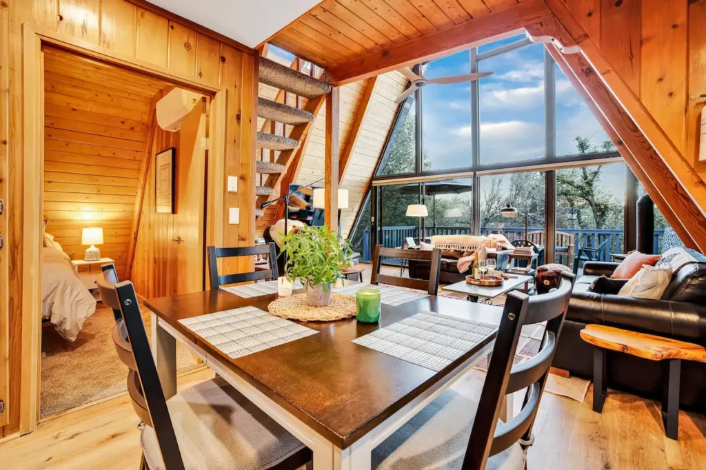Wooden dining table inside an A-frame cabin looking toward a living room with large glass windows