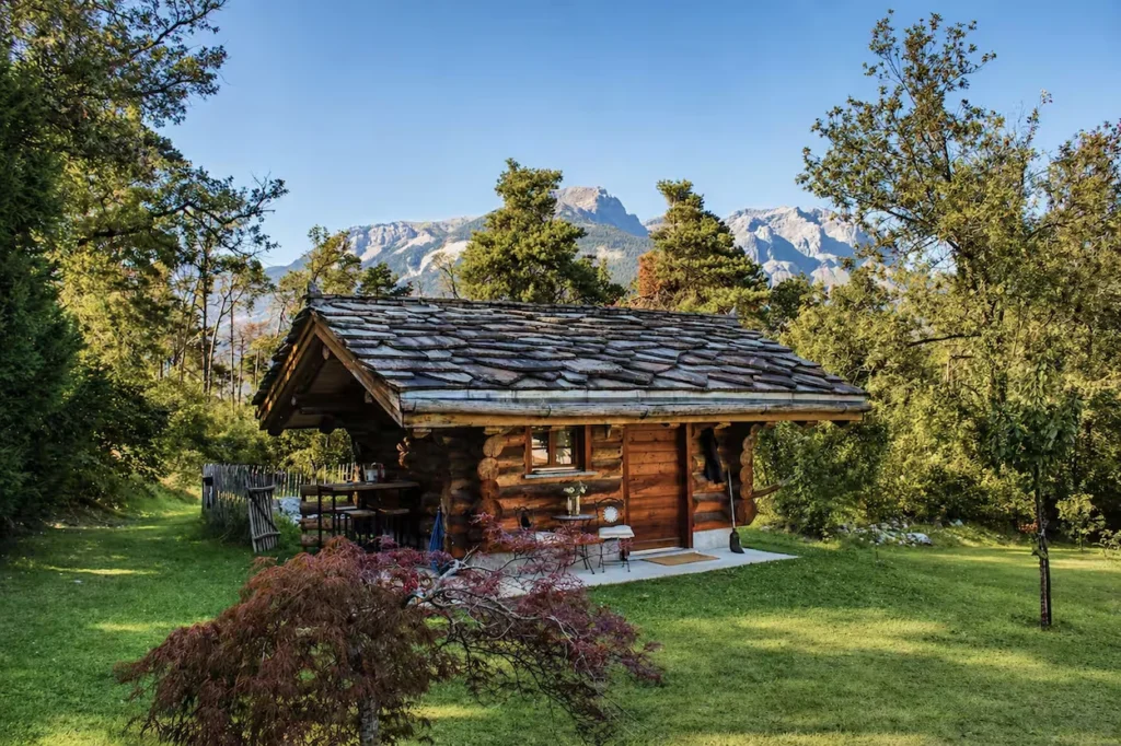 Rustic log cabin with a stone tile roof on a green lawn with rocky mountains in the background