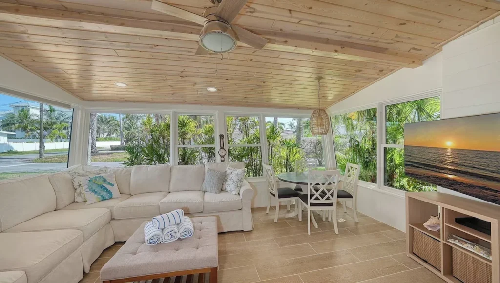 A sunlit beach cottage living room with a white sectional sofa, a wood-paneled ceiling, and a small dining set