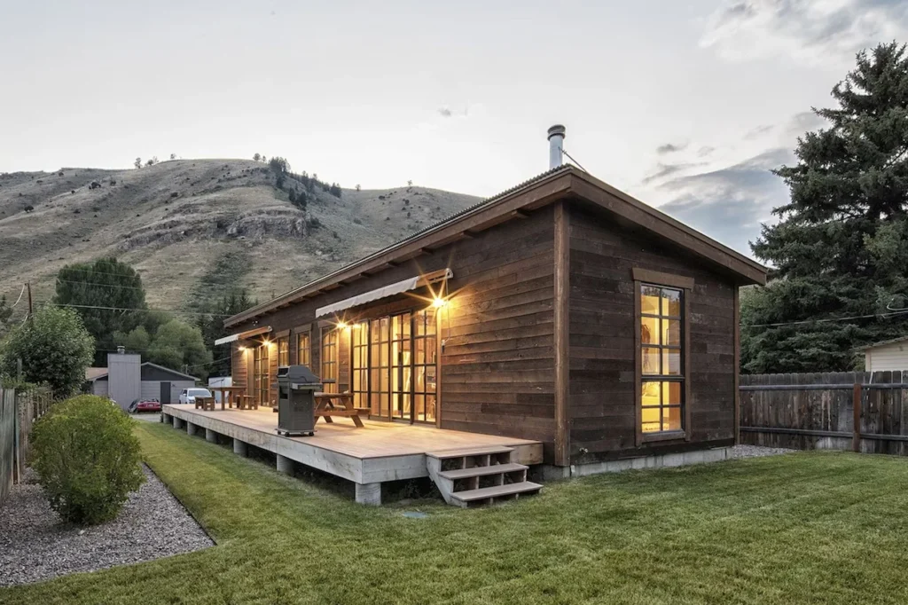 Modern wooden cabin with an illuminated long deck and grill under a rugged mountain backdrop