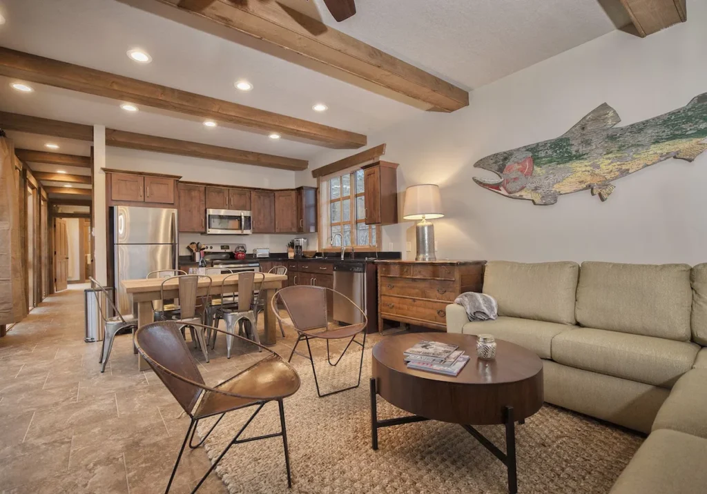 Open cabin interior with exposed wood beams, a tan sectional, metal dining chairs, and a large rustic fish wall art decoration