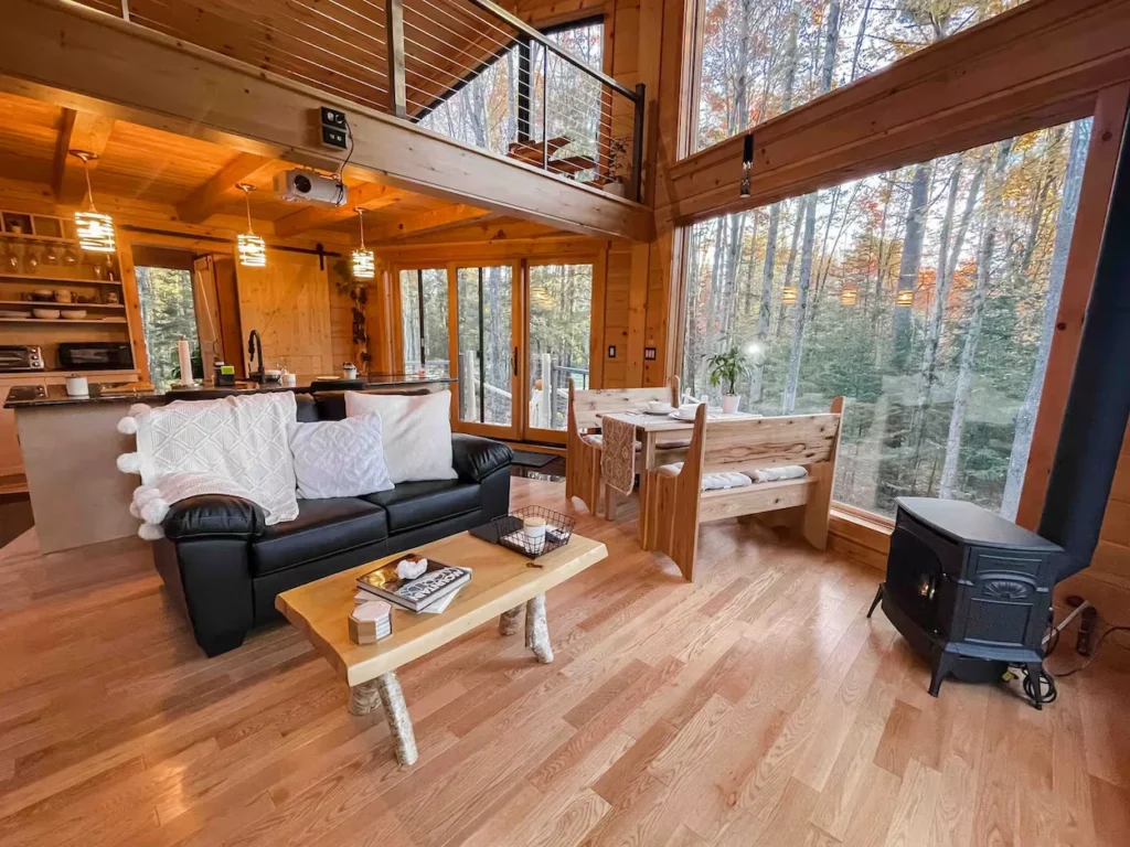 Cabin living room with floor-to-ceiling windows, black leather sofa, wood stove, and a dining booth