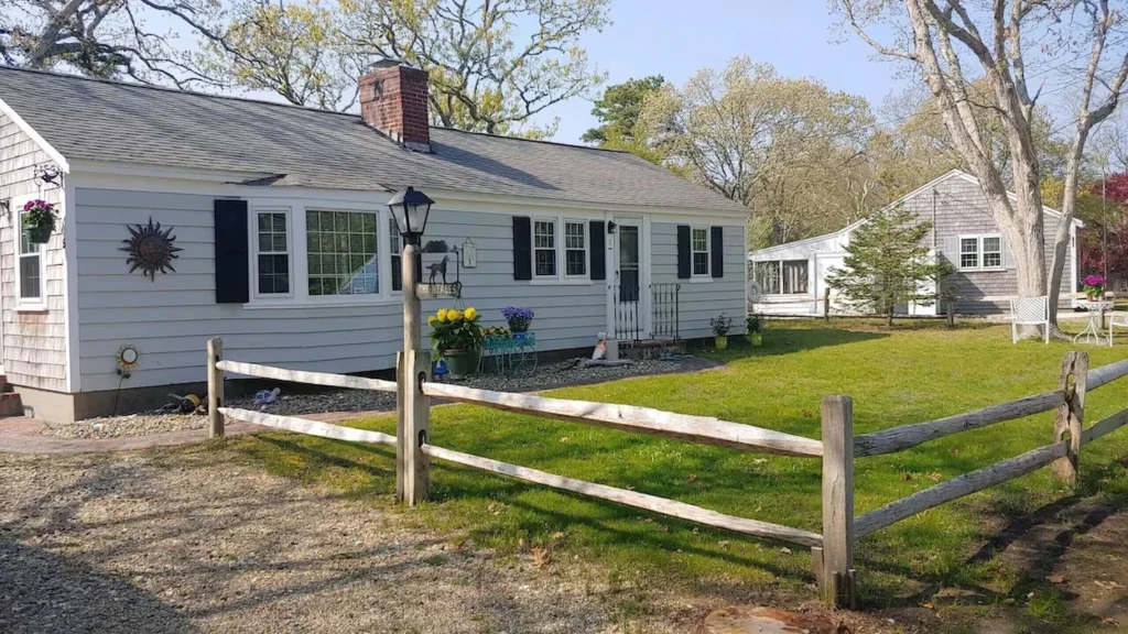 A gray single-story cottage with a wooden split-rail fence, gravel driveway, and green lawn