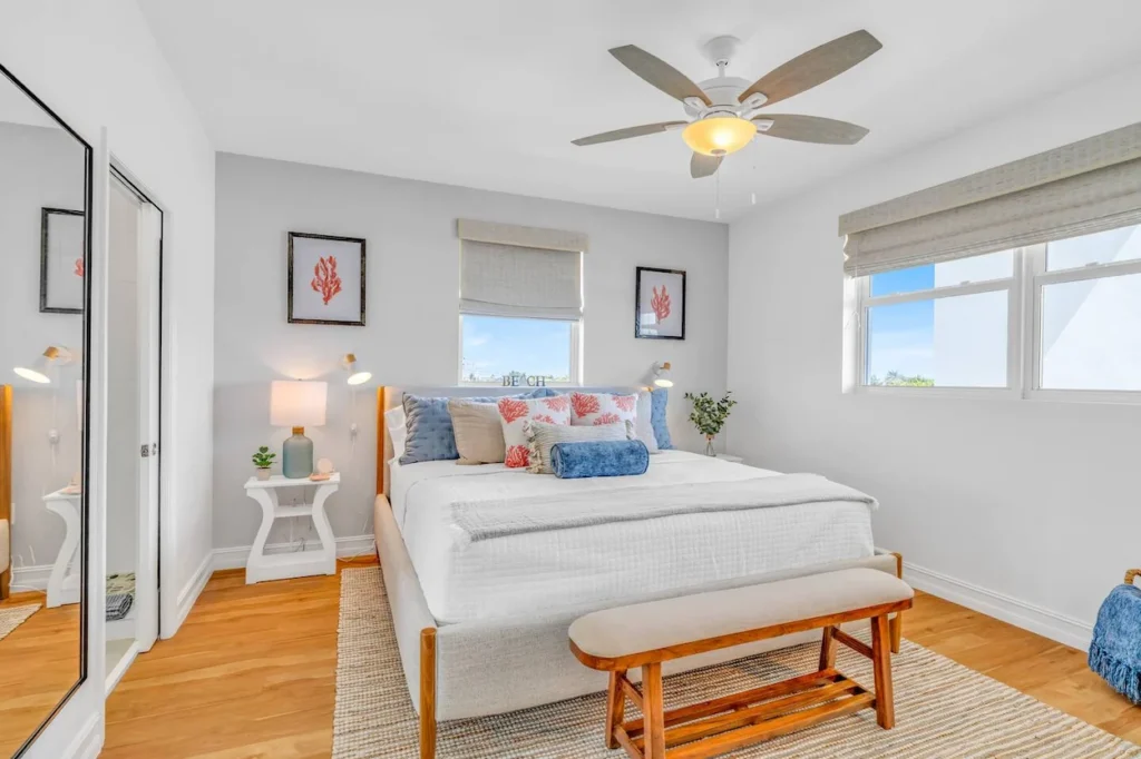 Bright beach house bedroom with a king bed, blue and coral accent pillows, a wooden bench, and a ceiling fan
