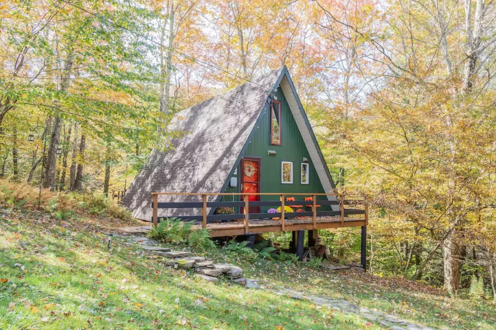 Green A-frame cabin with a wooden front deck surrounded by fall foliage