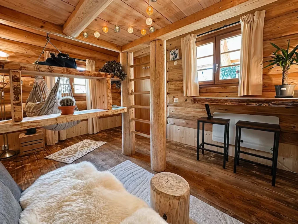 Rustic log cabin studio interior with string lights, a built-in ladder, a hammock chair, and bar stools