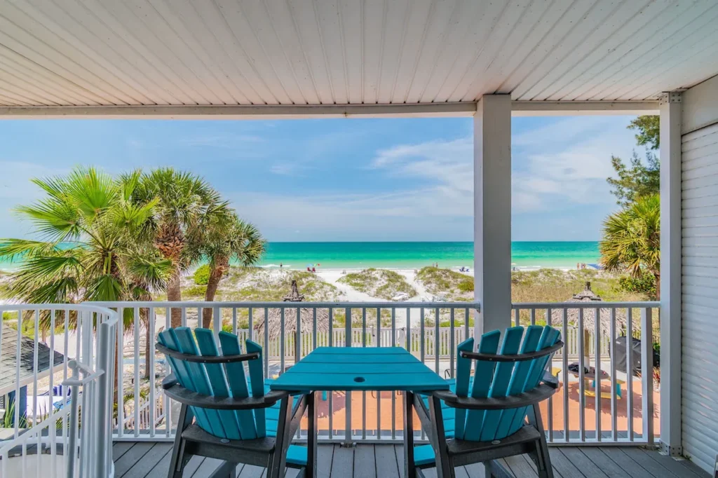 Two blue Adirondack chairs and a small table on a covered balcony facing a white sand beach and turquoise ocean