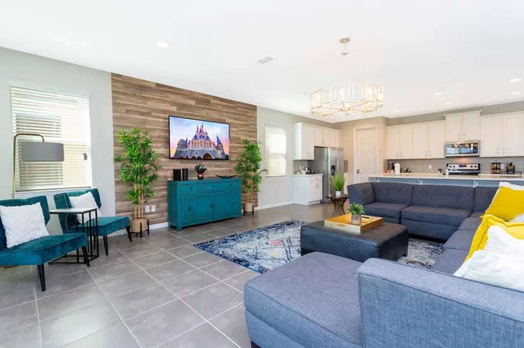 An open-concept living area with a large grey sectional sofa, tile floors, a teal accent credenza, and an adjoining white kitchen