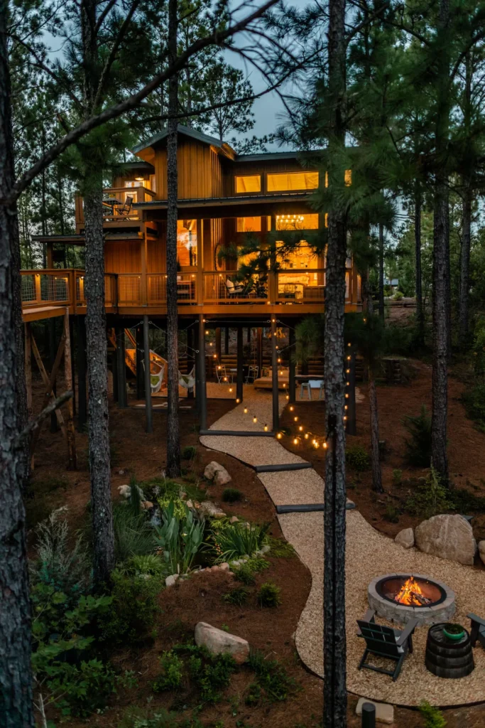 Two-story wooden treehouse cabin glowing with light at night with a stone fire pit area